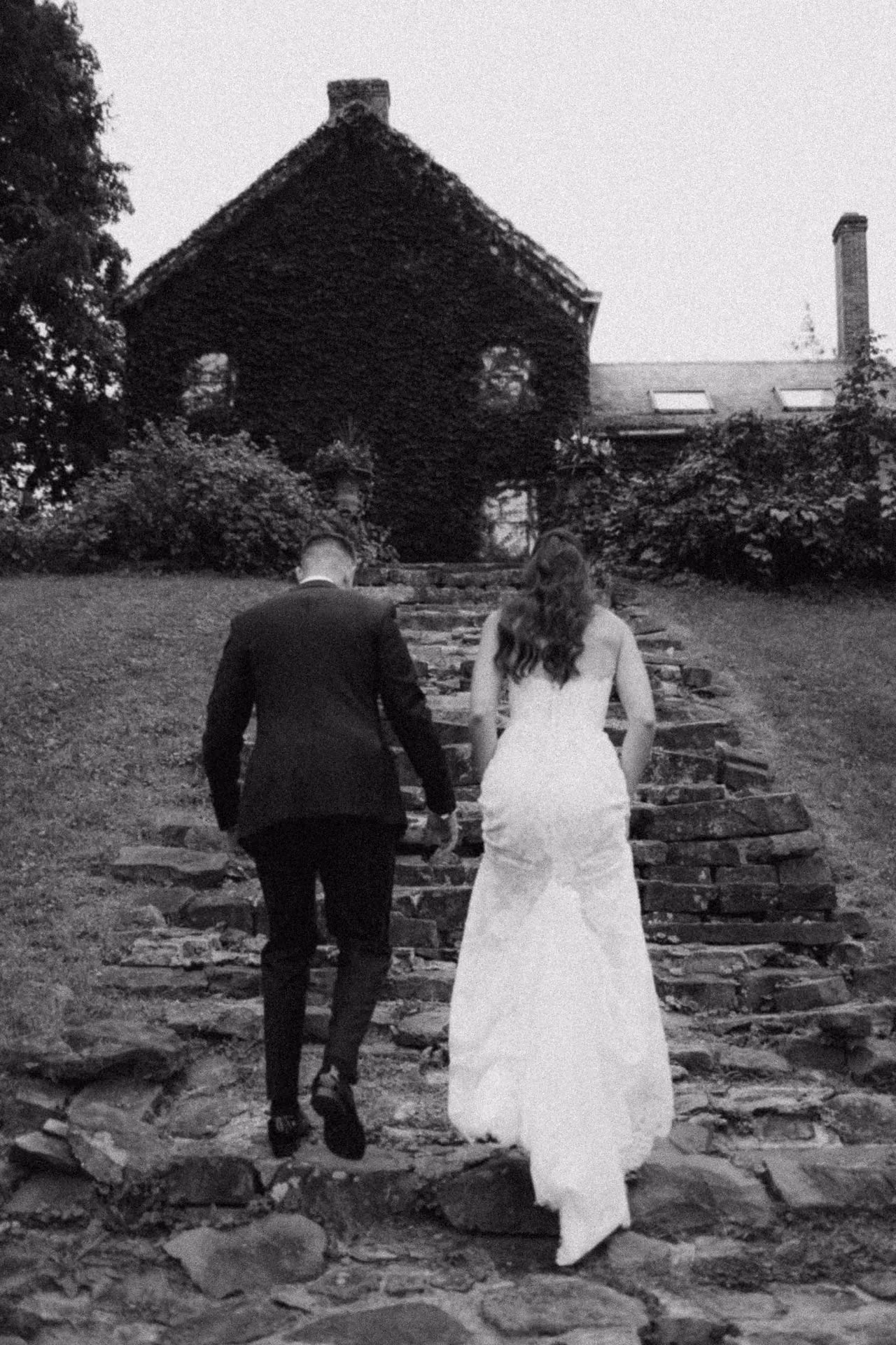 Black and white photograph of a bride and groom climbing stone steps toward an ivy-covered house at Windrift Hall