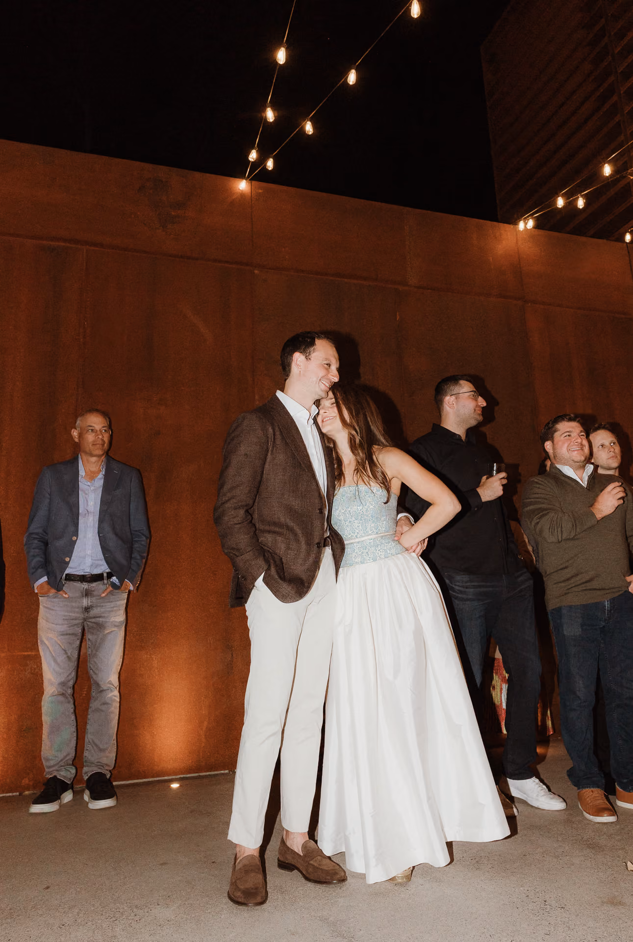 Couple smiling with friends against a lit wooden wall at a Camptown welcome party