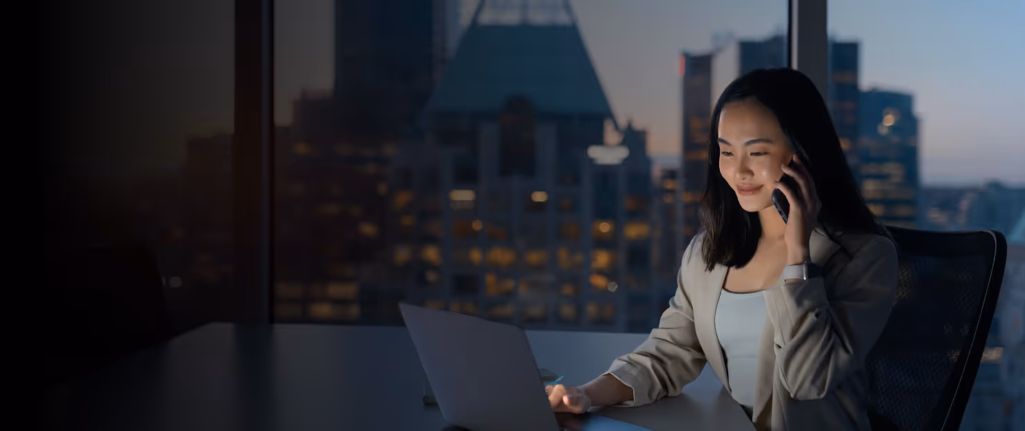 Woman in office jacket talking on phone while using laptop with city skyline at dusk outside window.
