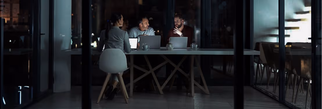 Three people with laptops engaged in a nighttime meeting in a modern glass-walled office.