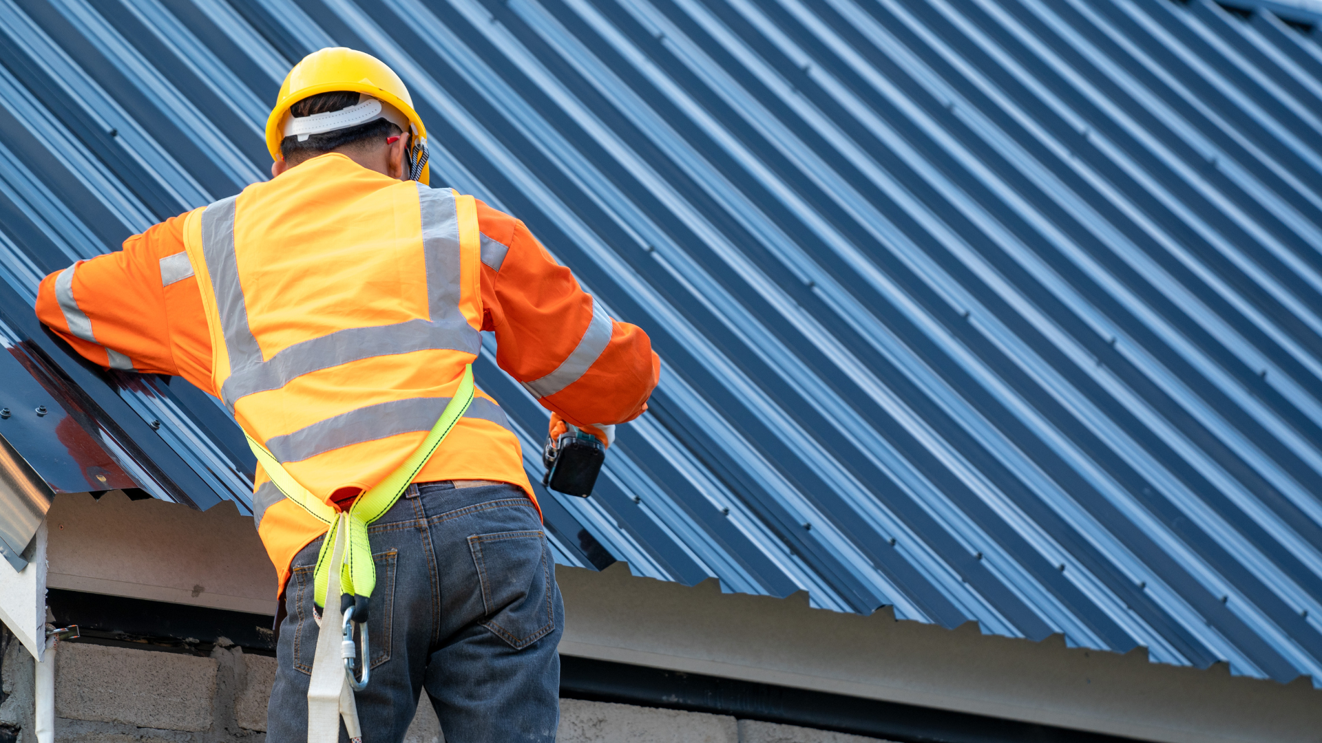 A roofer in orange reflective hear with a nail drill in hand drilling in a steel roofing system