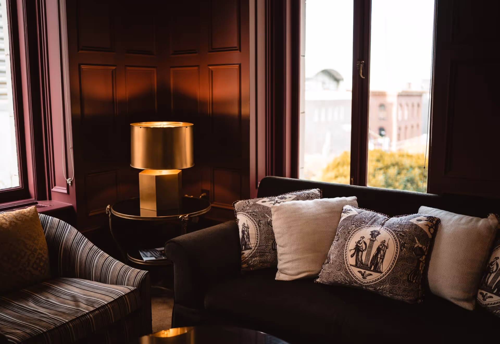 Indoor seating area with a striped armchair, sofa topped with decorative pillows, a gold table lamp, and a window view.