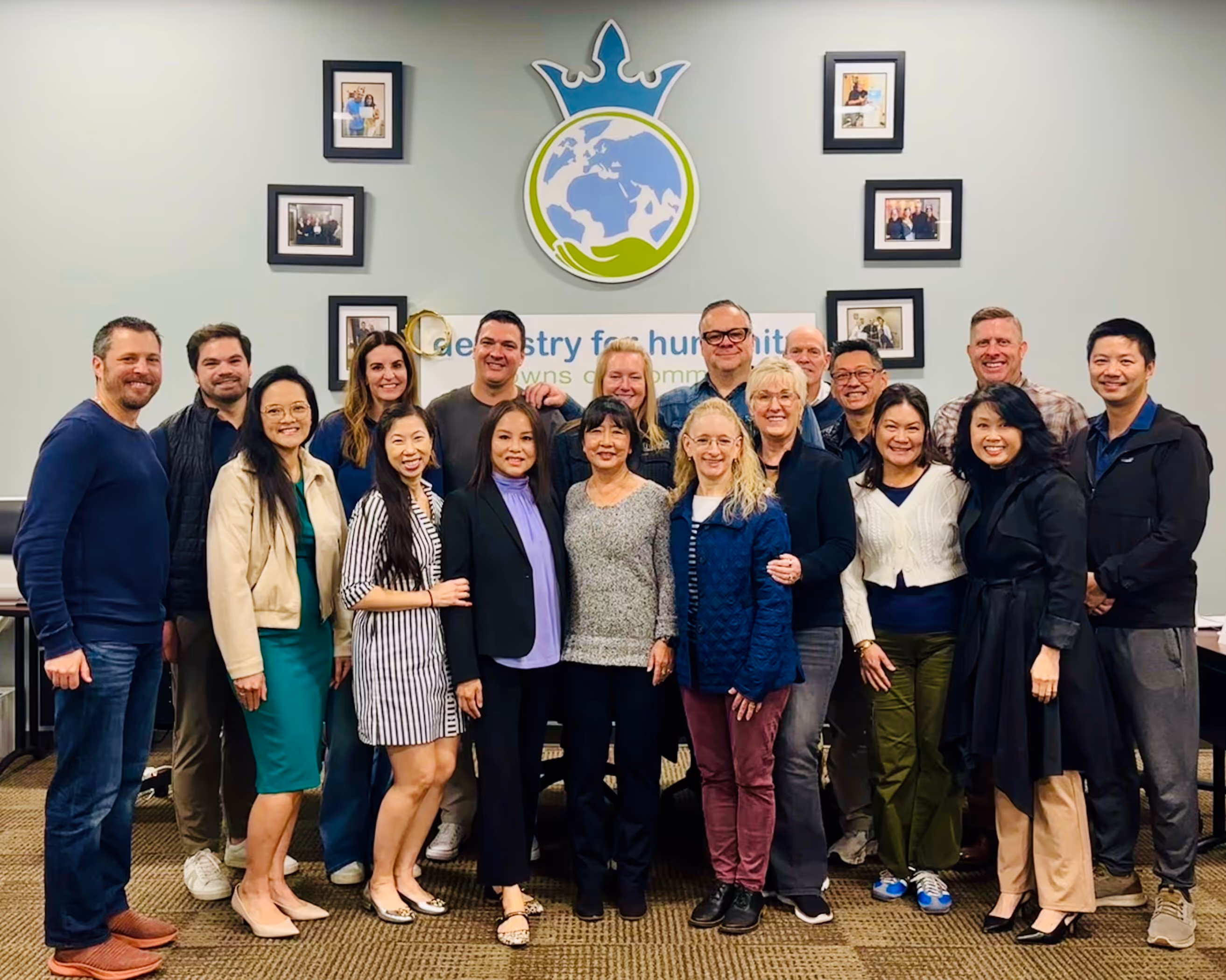 Group of 18 diverse adults standing indoors in front of a wall with a logo showing a globe with a crown and framed photos.