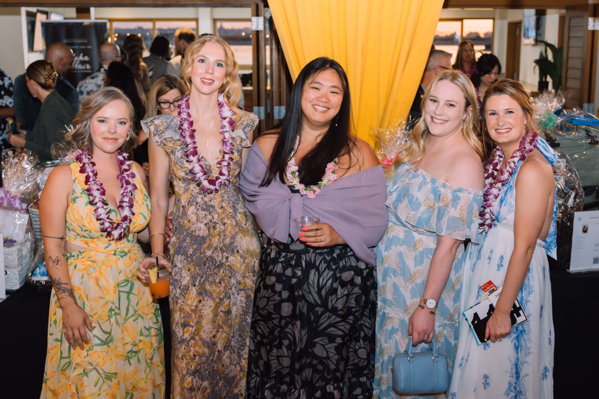 Five women wearing floral dresses and purple leis smiling and posing indoors at an event with yellow curtains and people in the background.