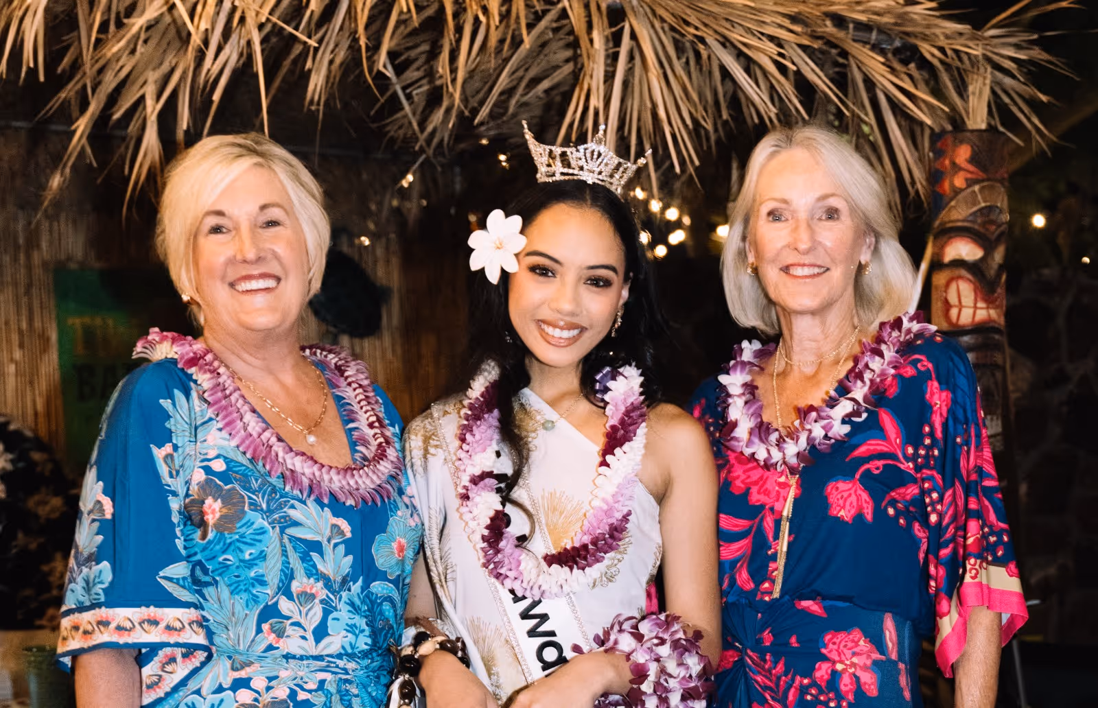 Three smiling women wearing floral dresses and leis, with the center woman wearing a crown and sash under a thatched roof.