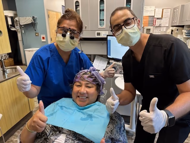 Two masked dental professionals giving thumbs-up behind a smiling seated patient wearing a blue bib.