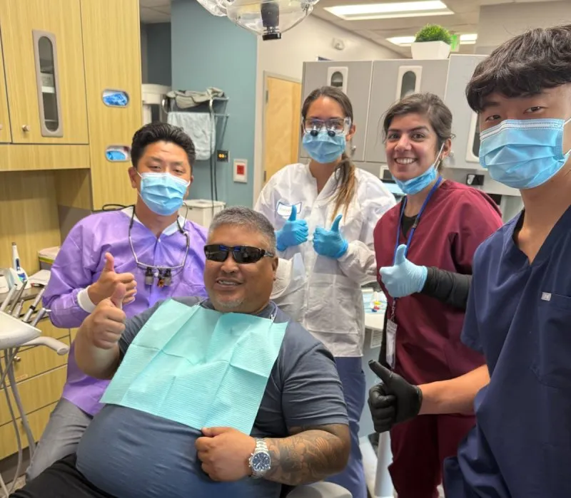 Smiling dental patient in chair giving thumbs up with four masked dental staff in clinic.