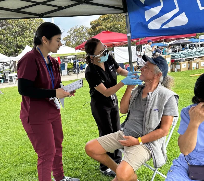 Healthcare worker in mask screens seated man outdoors while nurse with clipboard observes at event tent.
