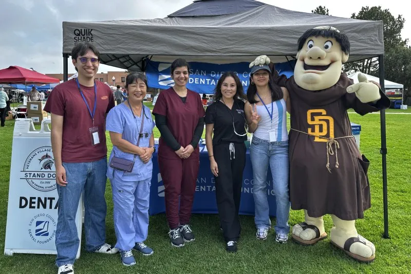 Six people, including one in a mascot costume, pose smiling under a tent at an outdoor dental event.