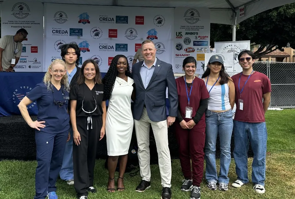Group of eight diverse people smiling and posing outdoors in front of event sponsor banners under a tent.