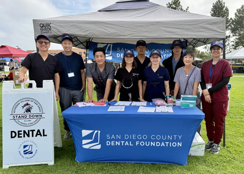 Group of dental professionals standing behind a San Diego County Dental Foundation booth with a white sign on grass.