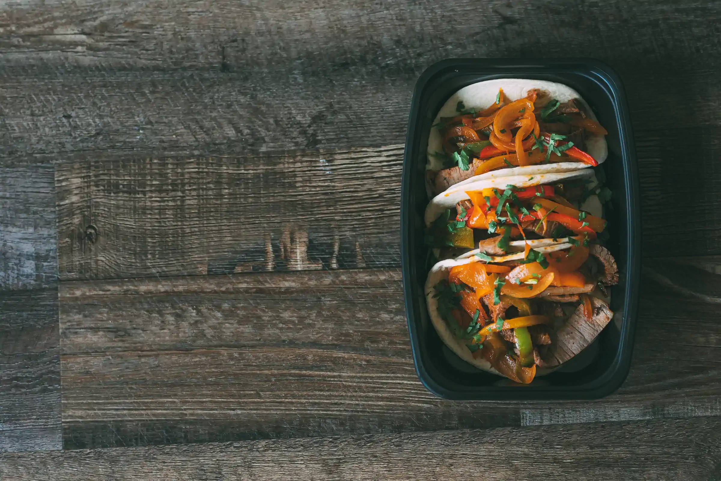 Three tacos filled with grilled meat, bell peppers, and garnished with fresh herbs, served in a black meal prep container on a rustic wooden table.
