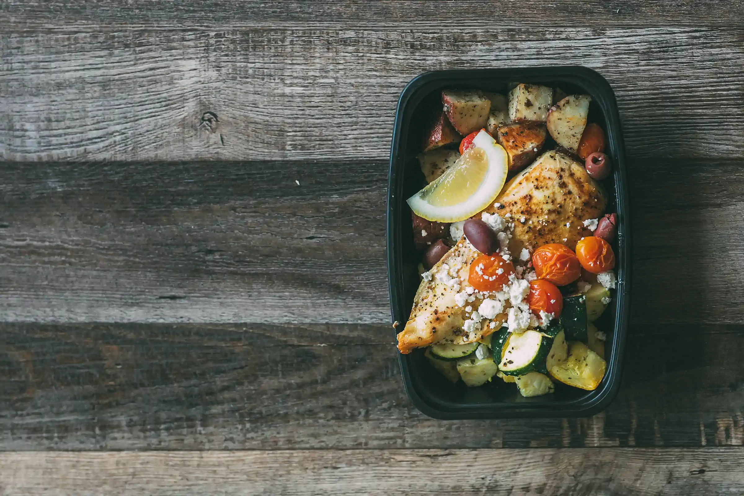 A black meal prep container holds a serving of seasoned chicken breast topped with cherry tomatoes, olives, crumbled feta cheese, and a lemon wedge. The dish is accompanied by roasted potatoes and zucchini. The container is placed on a rustic wooden surface.