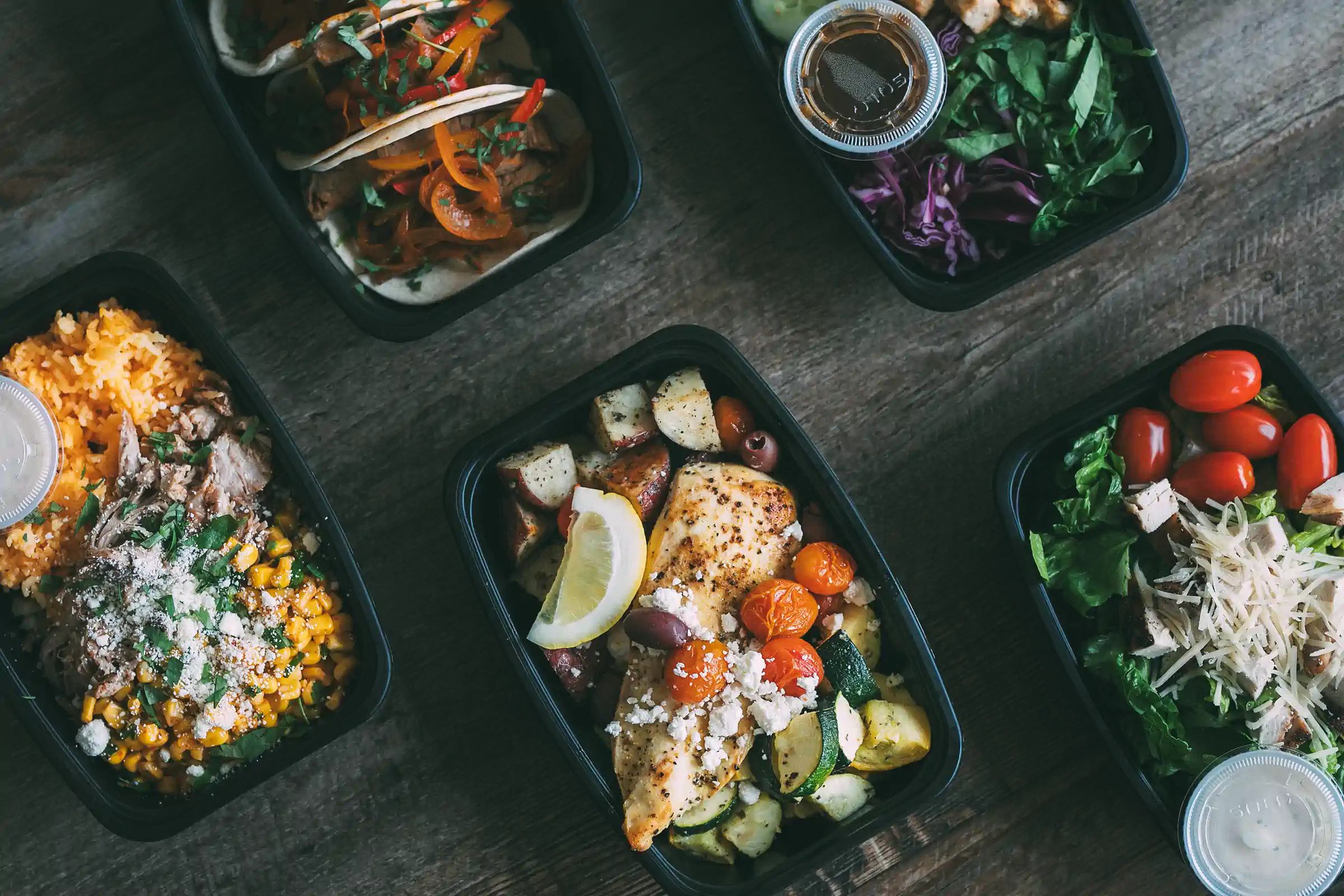 A variety of meal prep containers from Fed by Jed, featuring dishes such as grilled chicken with roasted vegetables, tacos with bell peppers, a salad with cherry tomatoes and shredded cheese, and a rice bowl with corn and shredded meat.
