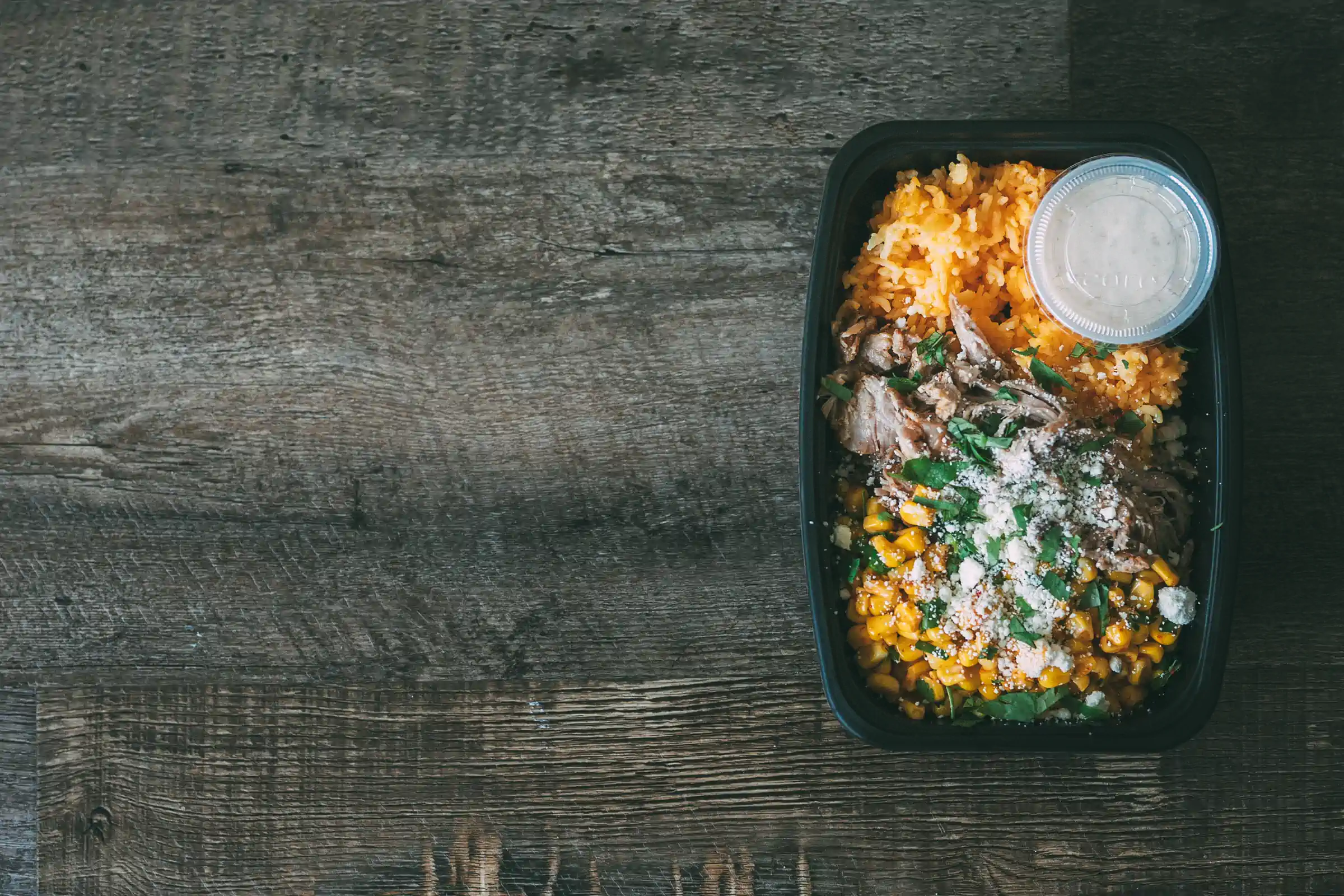 A black meal prep container from Fed by Jed containing shredded meat, corn, and rice, topped with herbs and cheese, with a small container of sauce on the side.