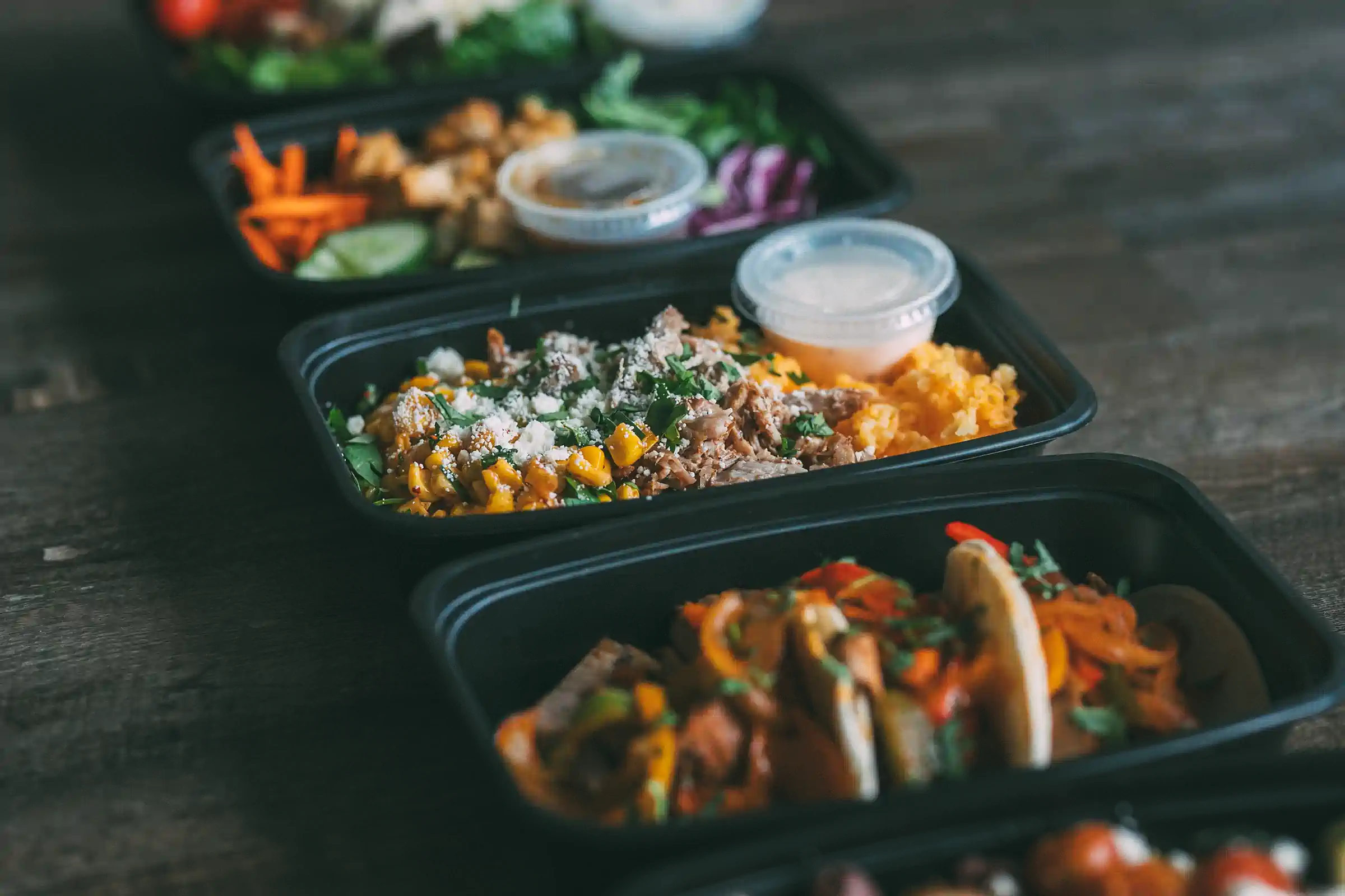 A row of black meal prep containers filled with various colorful dishes, including salads, tacos, and rice bowls, each with a small container of sauce on the side. The meals are part of Fed by Jed, a meal prep and delivery service in Milwaukee.