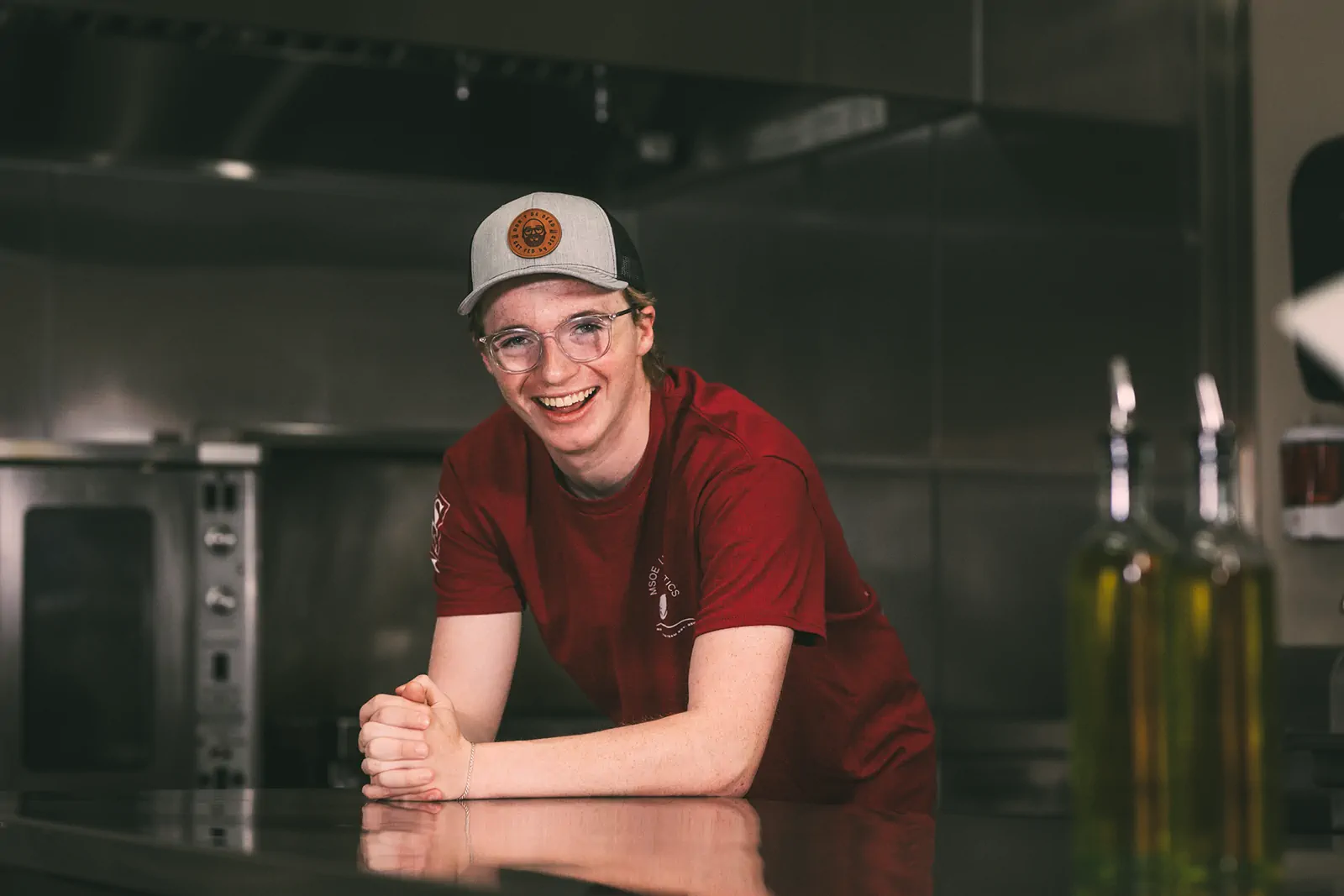 A portrait of Kian, Fed by Jed's delivery driver. A smiling young man wearing a gray cap and red "MSOE" t-shirt leans on a counter in a commercial kitchen.