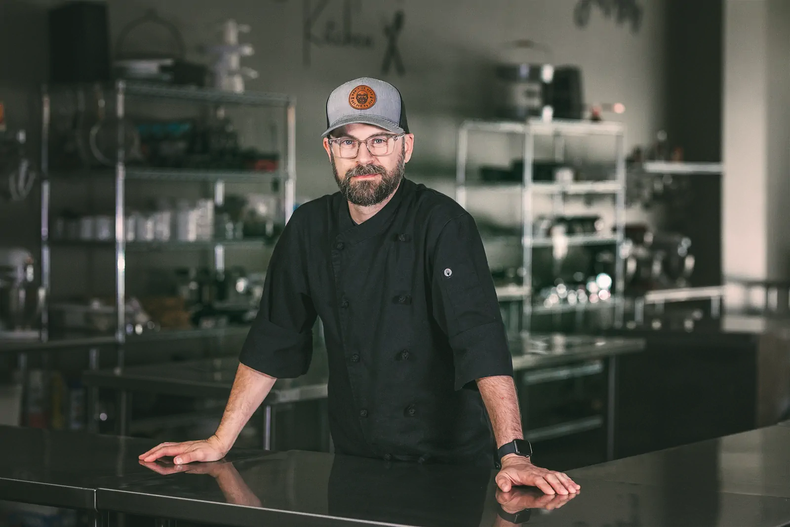 A bearded man wearing glasses, a black chef's coat, and a gray cap with a logo stands confidently in a commercial kitchen. He is leaning on a stainless steel counter with shelves of kitchen equipment in the background. This is Jed, owner of the meal prep and delivery service Fed by Jed in Milwaukee.