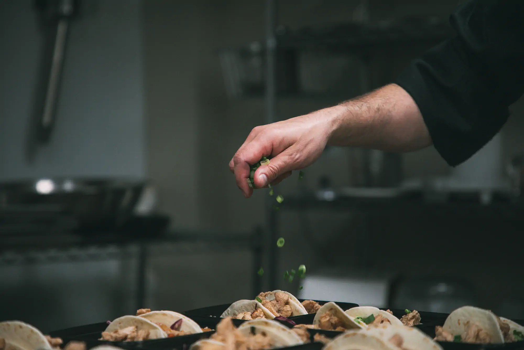 A chef's hand sprinkles chopped green onions over a tray of prepared tacos in a kitchen.