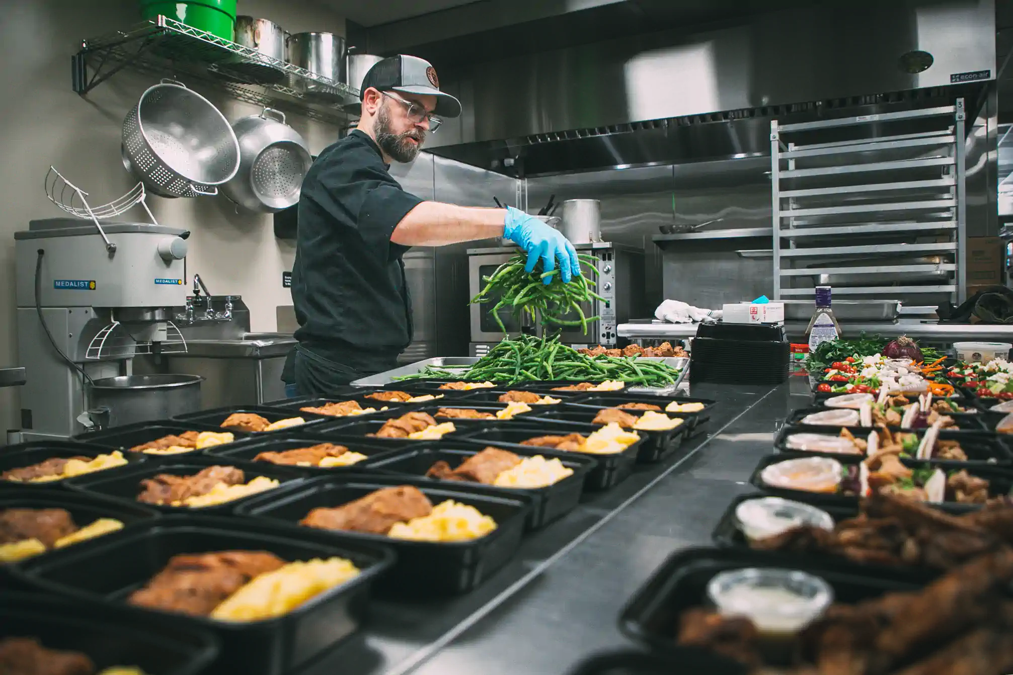 A chef in a commercial kitchen prepares meal trays, adding green beans to each tray. The kitchen is equipped with industrial appliances, including a Medalist mixer and various cooking utensils. The trays contain portions of food, ready for delivery by Fed by Jed, a meal prep and delivery service in Milwaukee.