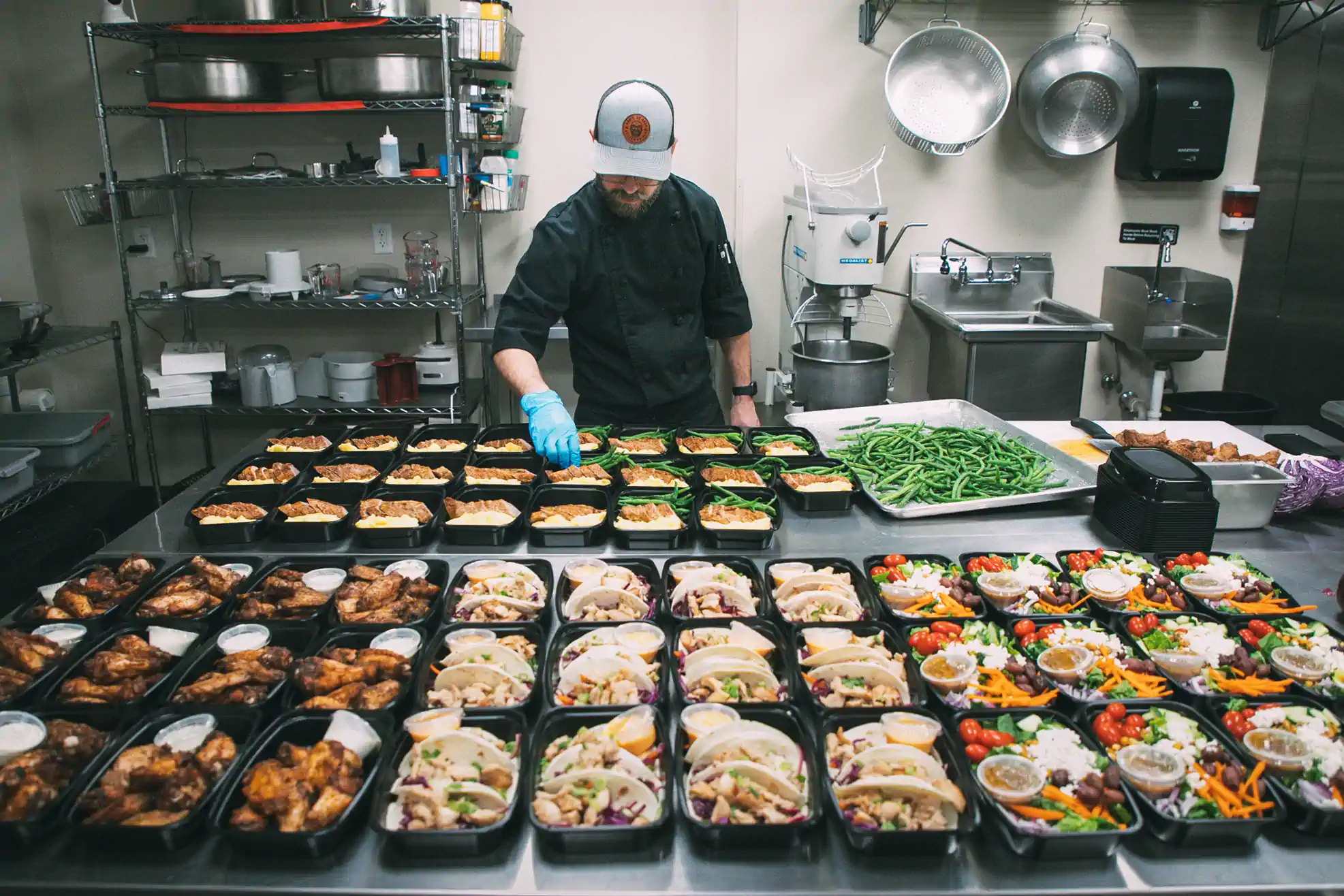 A chef in a commercial kitchen prepares multiple meal prep containers filled with various dishes, including green beans, chicken wings, tacos, and salads. Shelves with kitchen equipment and ingredients are visible in the background.