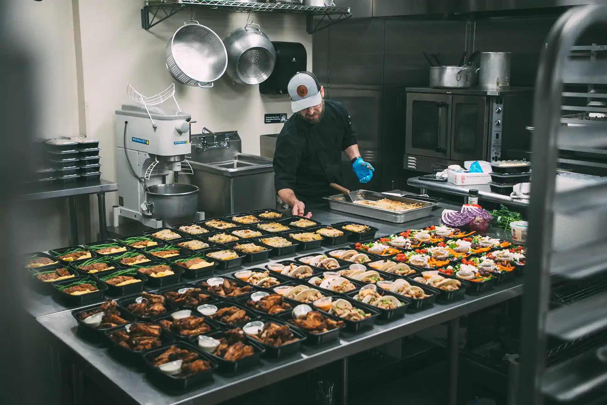 A chef in a commercial kitchen prepares multiple meal prep containers filled with various dishes. The containers are neatly arranged on a large stainless steel table. The chef wears a black uniform, a cap, and blue gloves. The kitchen is equipped with industrial appliances, including a mixer labeled "MEDALIST" and ovens in the background.