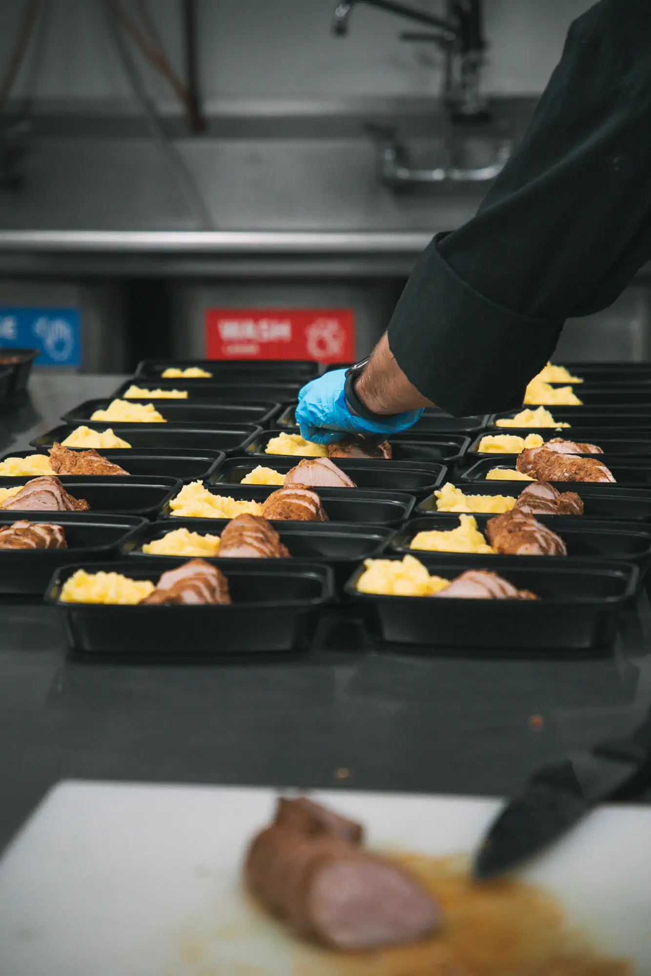 A person wearing a black uniform and blue gloves is preparing meal trays with sliced meat and mashed potatoes in a commercial kitchen. The trays are neatly arranged in rows on a countertop.