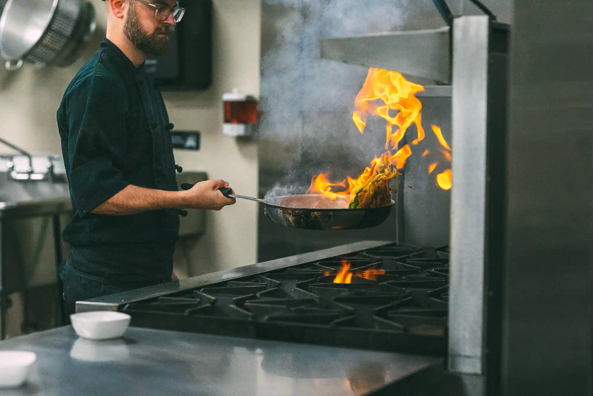 A chef in a professional kitchen is flambéing vegetables in a pan over a gas stove, with flames rising high. The chef is wearing a dark uniform and glasses.