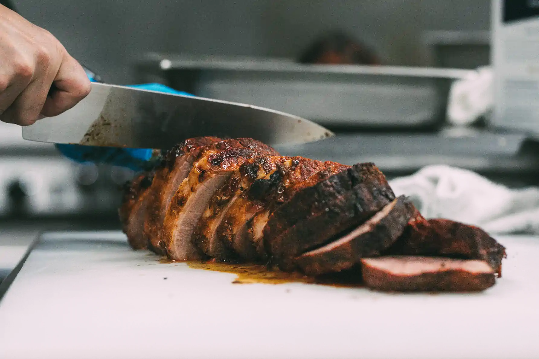 A person slices a seasoned, cooked meat roast on a white cutting board in a kitchen.