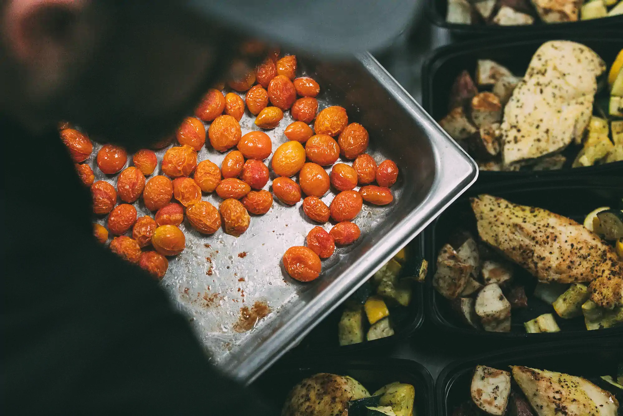 A person is preparing meal prep containers with roasted cherry tomatoes, seasoned chicken breast, and mixed roasted vegetables.