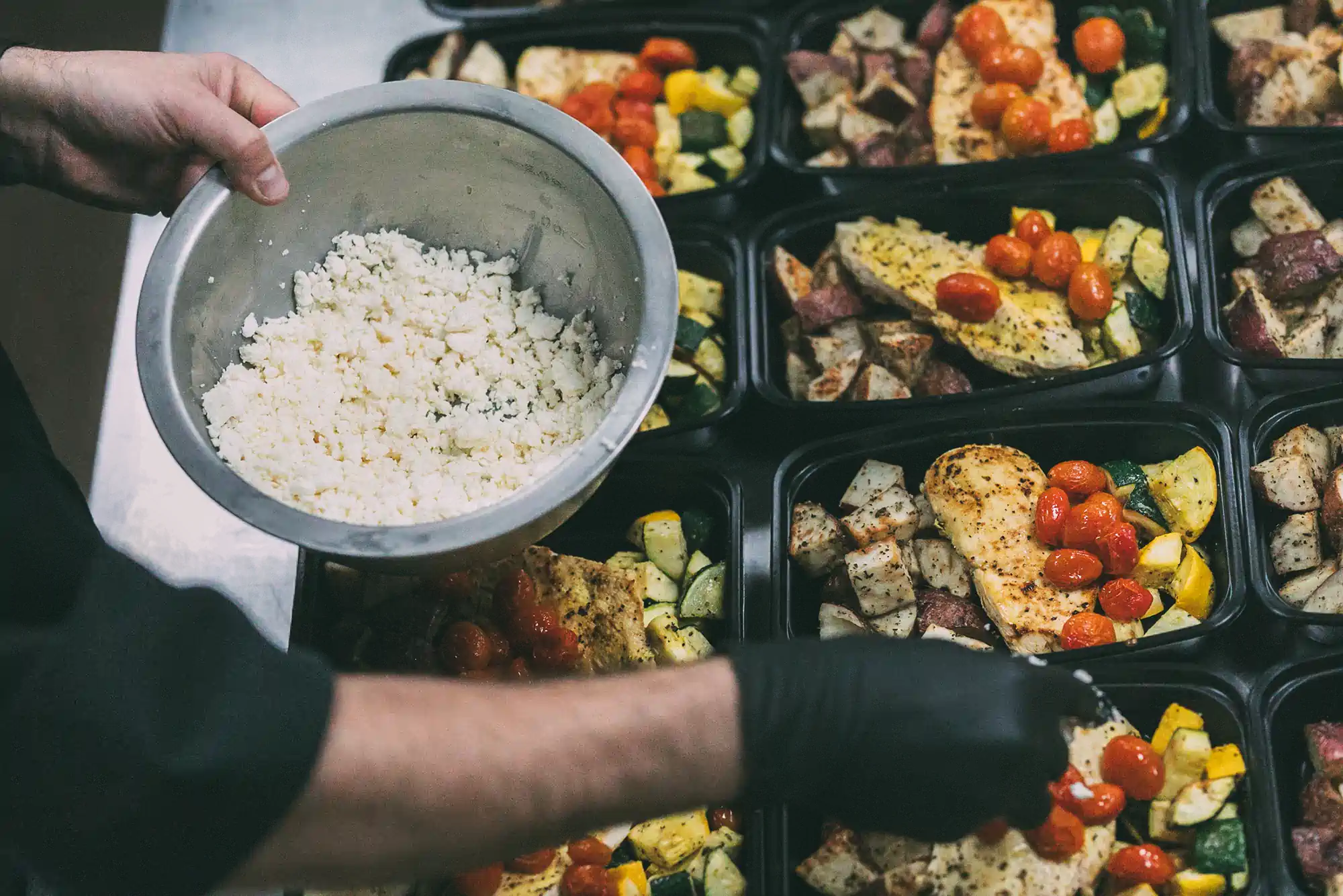 A person wearing black gloves is preparing meal prep containers with seasoned chicken, roasted potatoes, zucchini, and cherry tomatoes. They are holding a bowl of crumbled cheese, ready to add it to the meals. The containers are neatly arranged on a countertop.