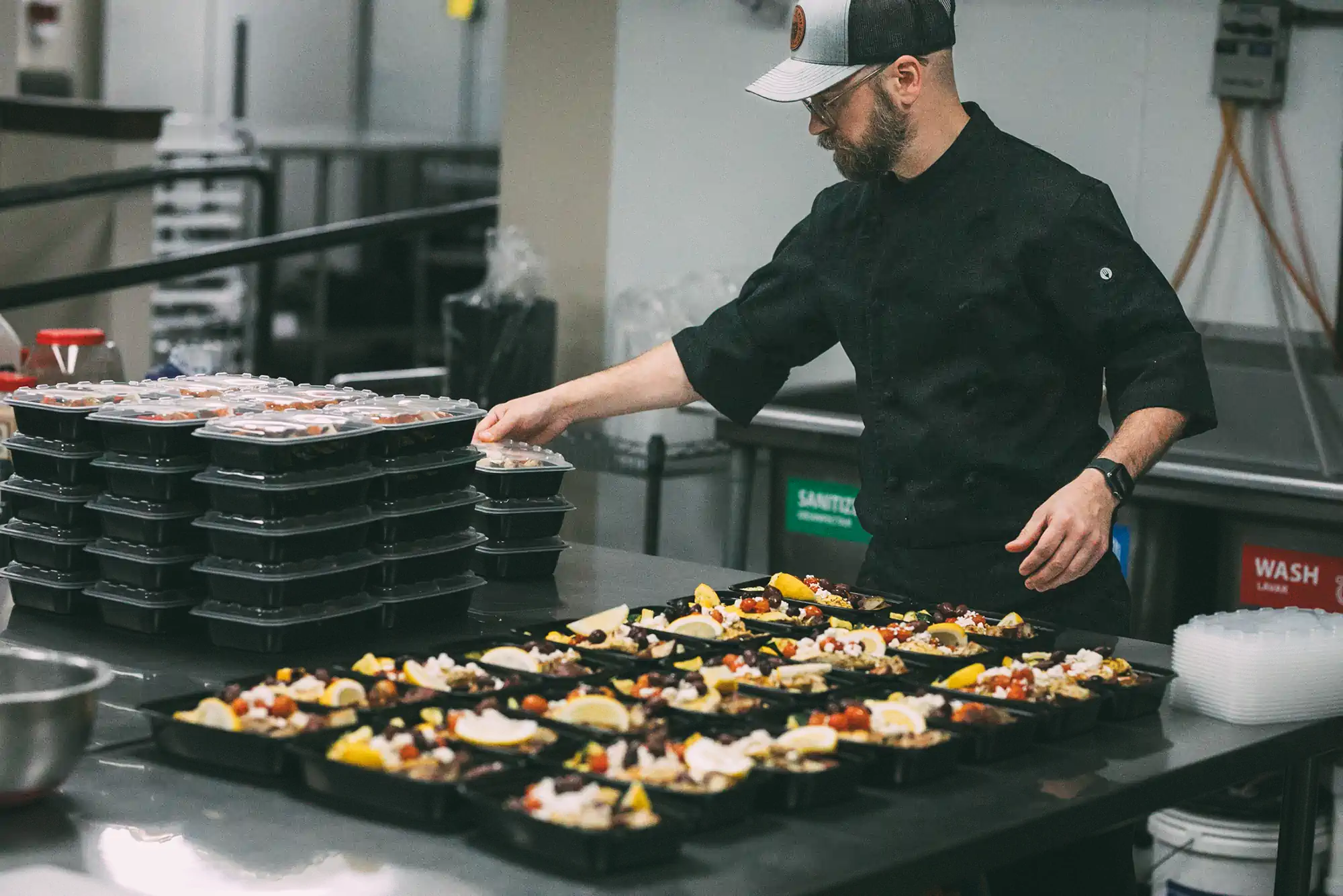 A man in a black chef's uniform and a cap is preparing meal trays in a commercial kitchen. He is stacking covered meal trays while other trays with colorful food are laid out on the counter. The kitchen has a clean and organized setup with labeled bins in the background.