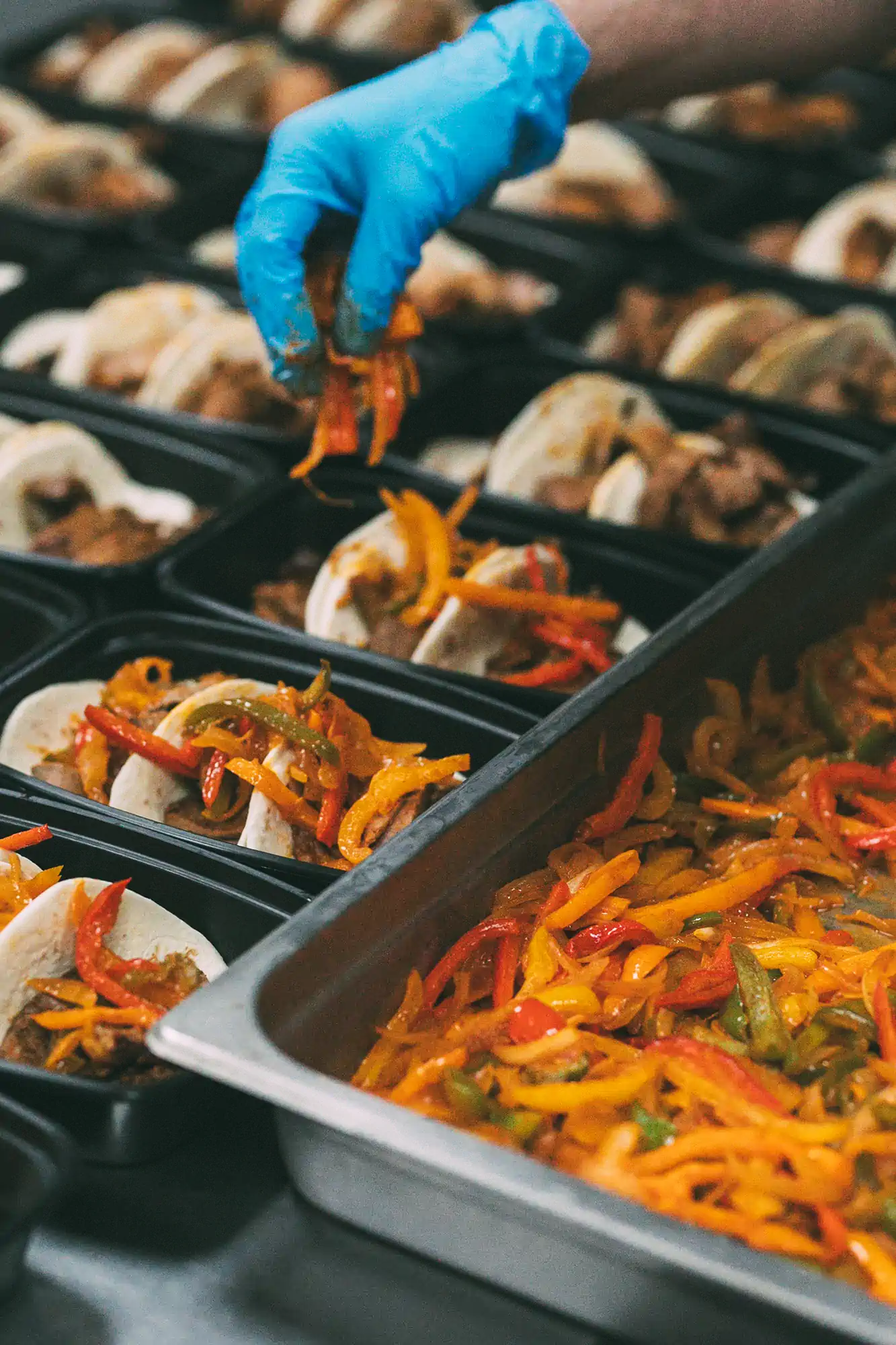 A gloved hand places cooked bell peppers into meal prep containers filled with tacos, part of the Fed by Jed meal prep and delivery service in Milwaukee.