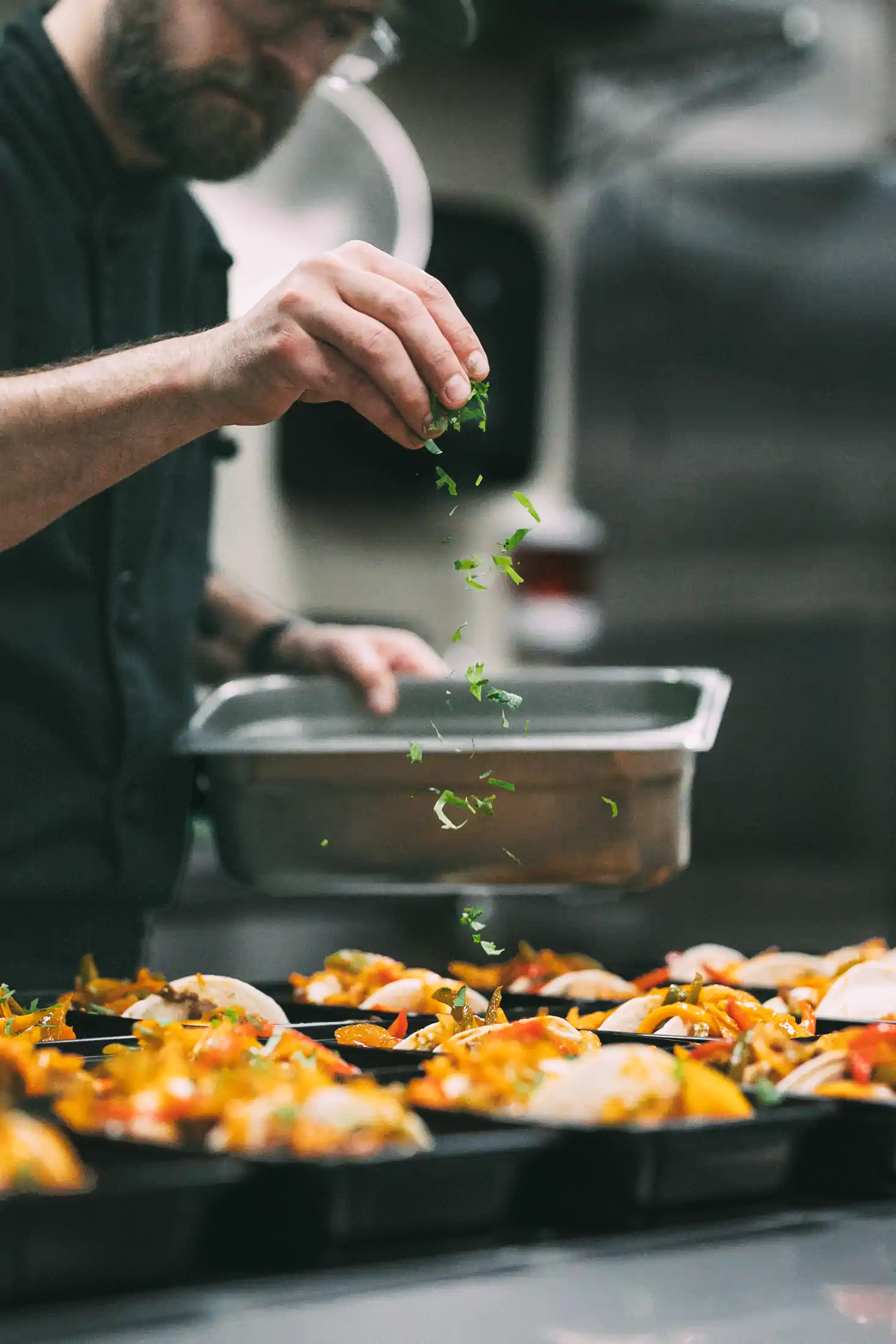 A chef sprinkles fresh herbs over trays of colorful, prepared meals in a kitchen.