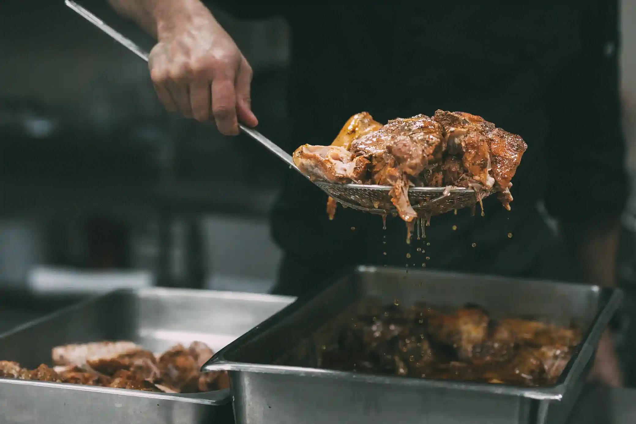 A chef is using a slotted spoon to transfer cooked meat from one metal tray to another in a kitchen. The meat is dripping with juices.