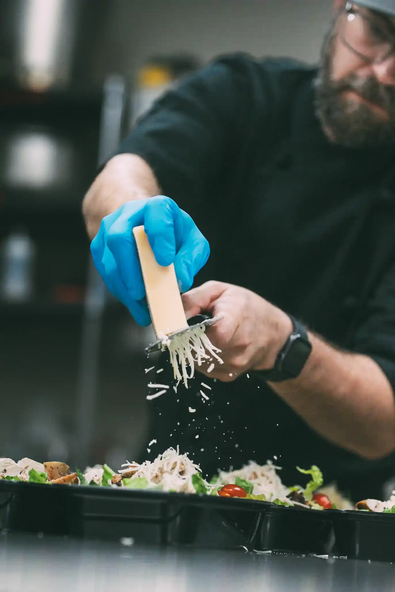 A chef wearing blue gloves grates cheese over prepared meal trays filled with salad ingredients, including cherry tomatoes and lettuce.