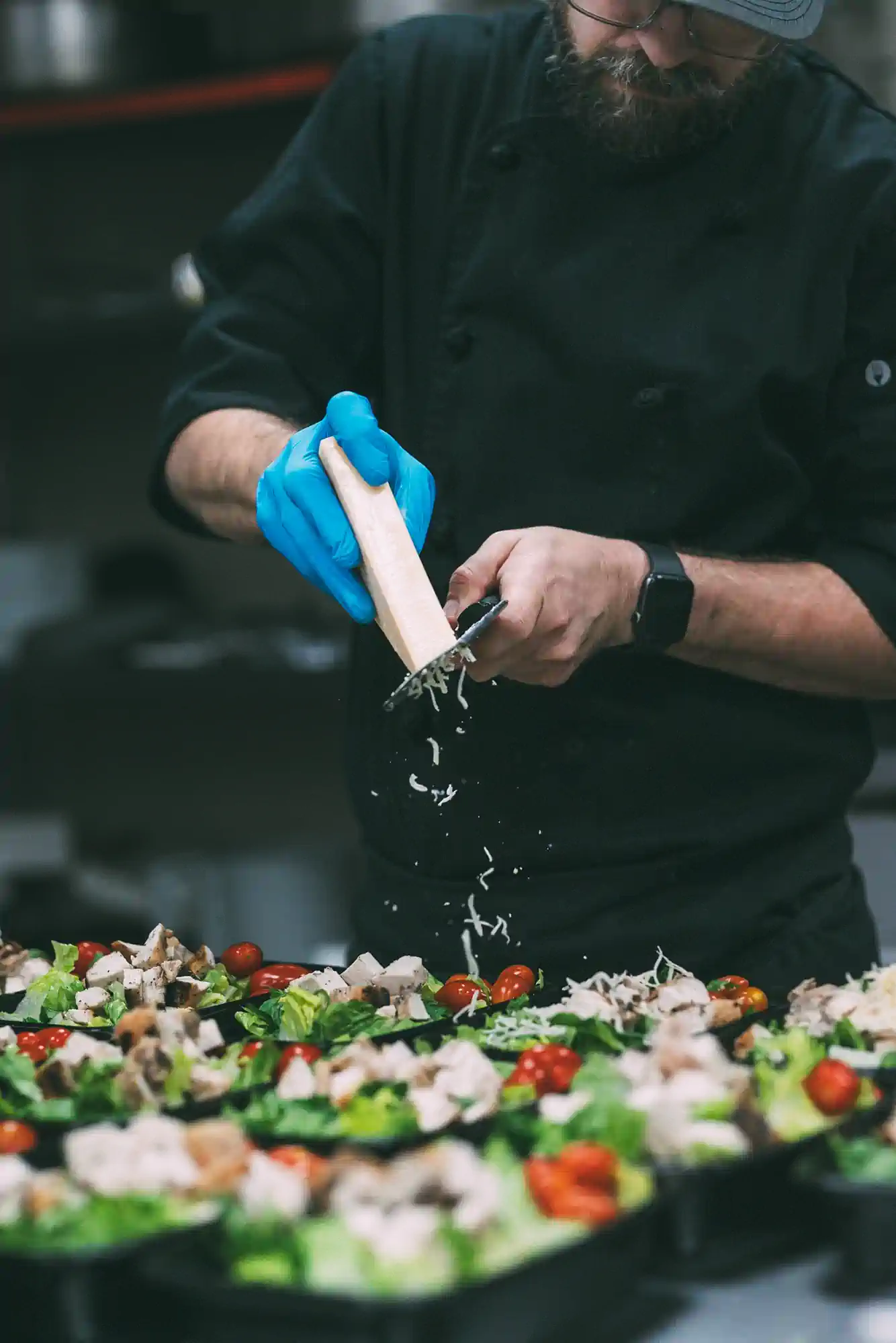 A chef wearing blue gloves and a black uniform grates cheese over several prepared salads with cherry tomatoes, lettuce, and chicken.