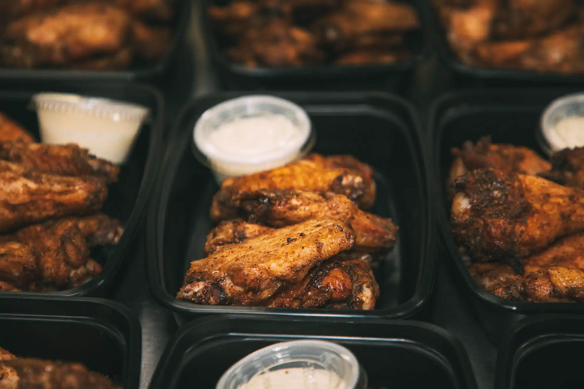 Black meal prep containers filled with seasoned chicken wings and small cups of dipping sauce, prepared by Fed by Jed, a meal prep and delivery service in Milwaukee.