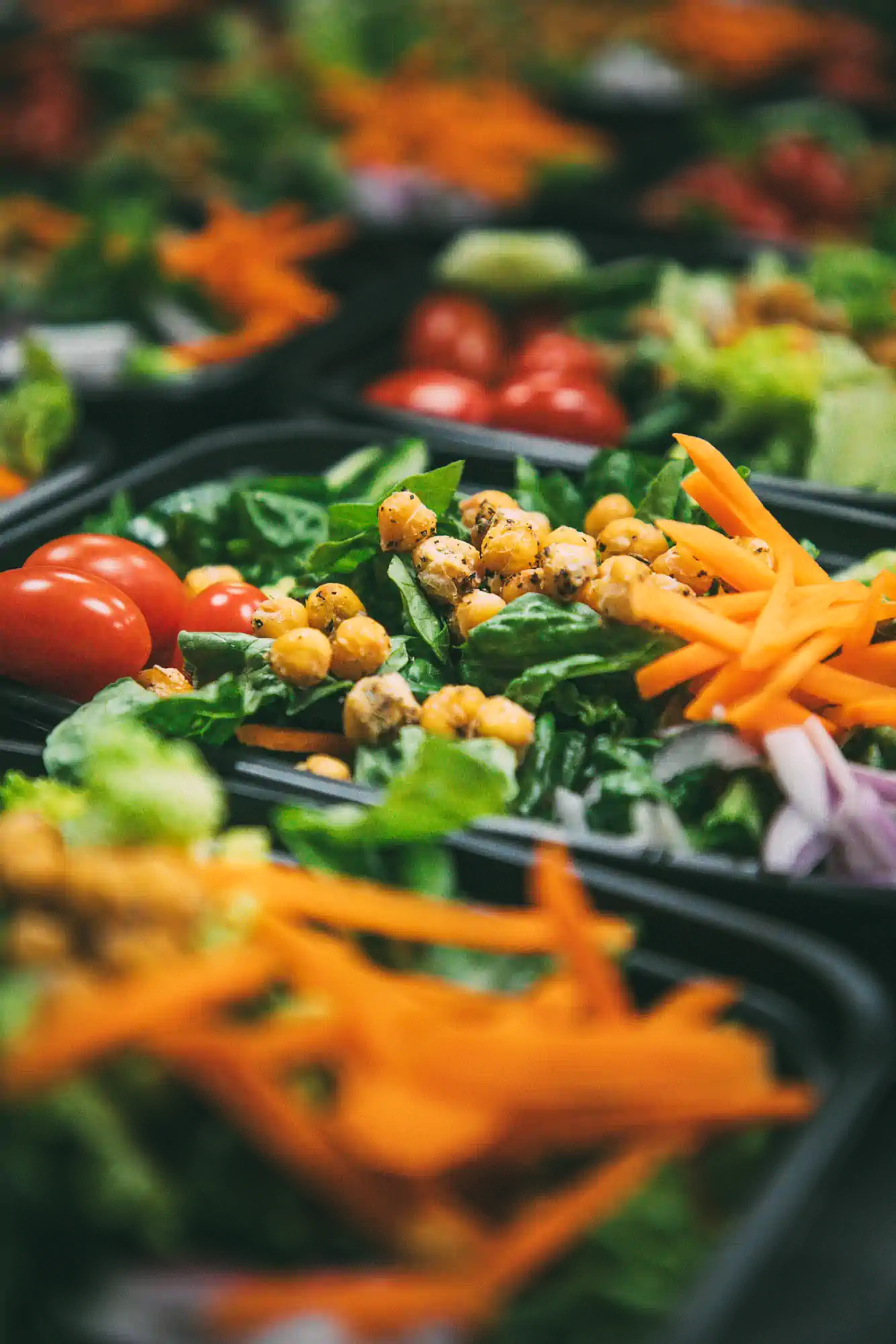 A close-up of a fresh salad in a black container, featuring cherry tomatoes, chickpeas, shredded carrots, spinach, and red onions. Multiple similar salads are visible in the background.