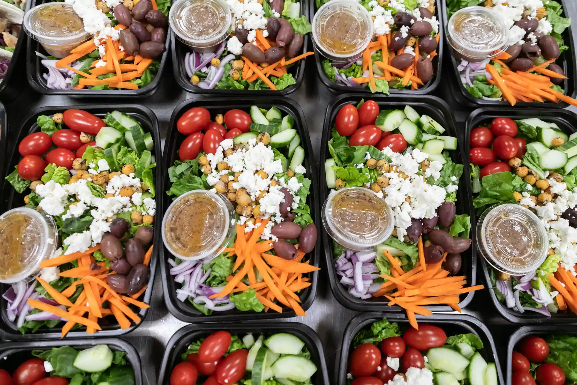 A set of meal prep containers filled with fresh salads, including cherry tomatoes, cucumbers, carrots, red onions, chickpeas, Kalamata olives, feta cheese, and a small container of dressing. The salads are neatly arranged and ready for delivery by Fed by Jed, a meal prep and delivery service in Milwaukee.