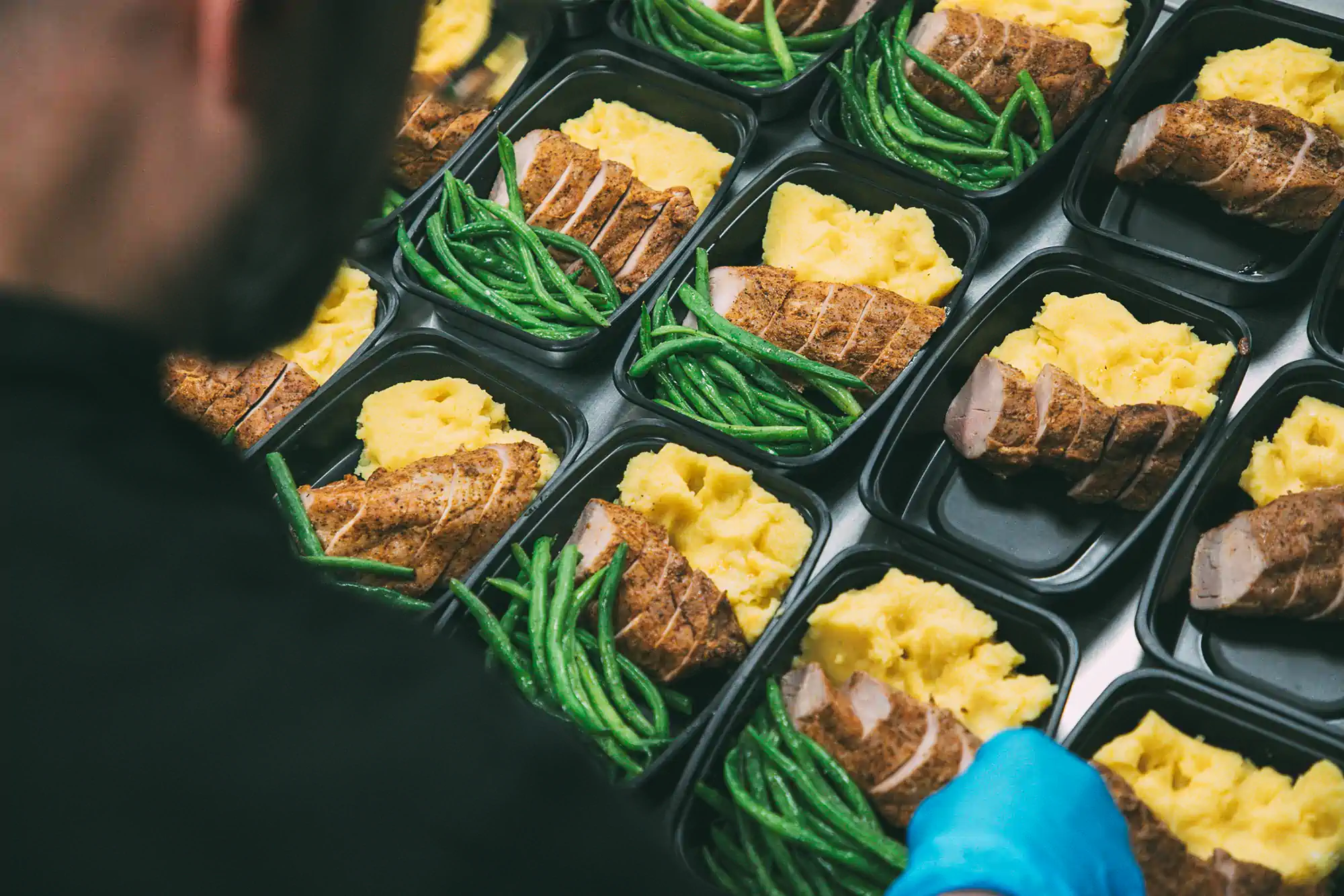 An over the shoulder shot of Jed wearing a blue glove, preparing meals containing sliced seasoned meat, mashed potatoes, and green beans. The trays are neatly arranged in rows, ready for delivery.