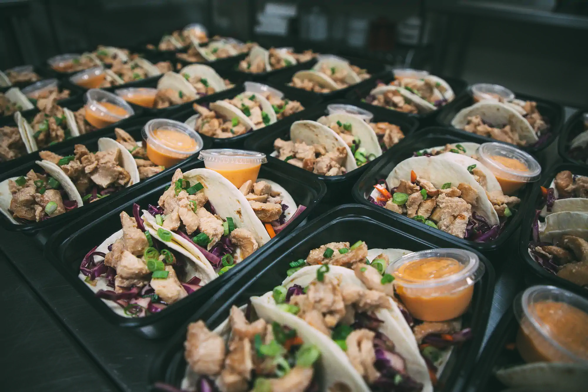 Rows of meal prep containers filled with tacos, topped with diced green onions and accompanied by small containers of orange sauce. The tacos are prepared by Fed by Jed, a meal prep and delivery service in Milwaukee.