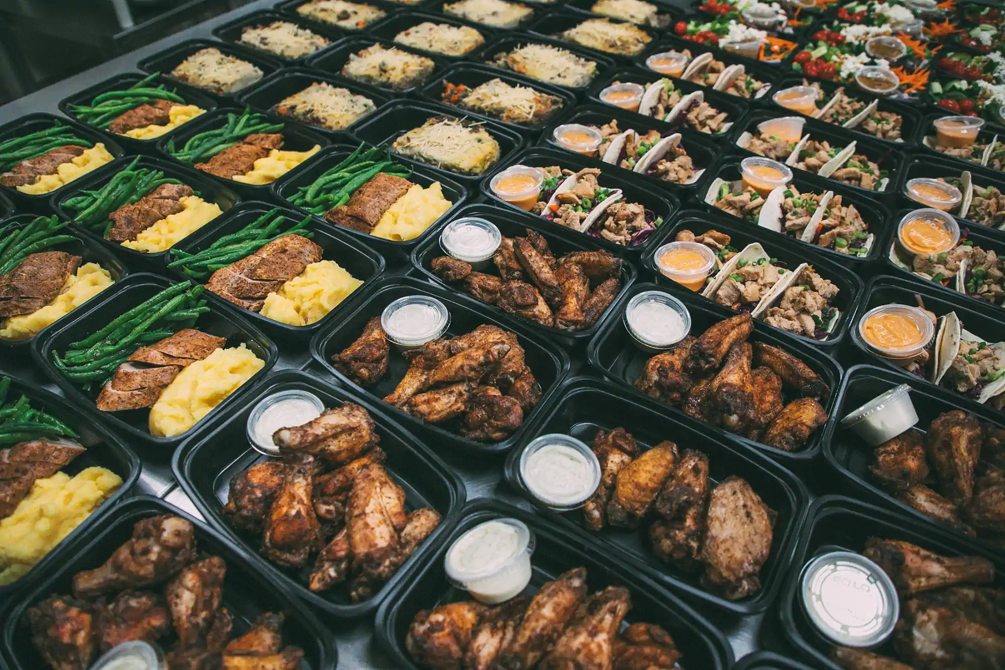 A variety of meal prep containers filled with different dishes, including green beans, mashed potatoes, sliced meat, chicken wings, tacos, and dipping sauces, arranged in rows on a table.