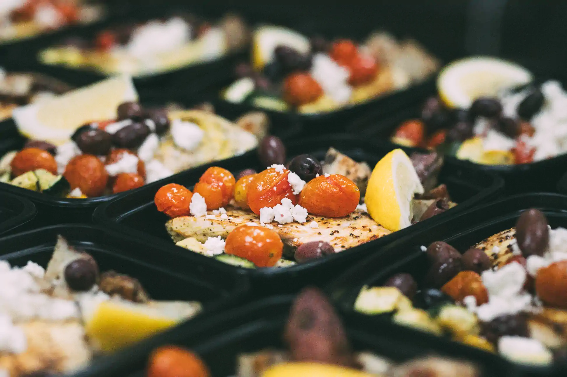 Close-up of meal prep containers from Fed by Jed, featuring grilled chicken topped with roasted cherry tomatoes, olives, feta cheese, and lemon wedges.
