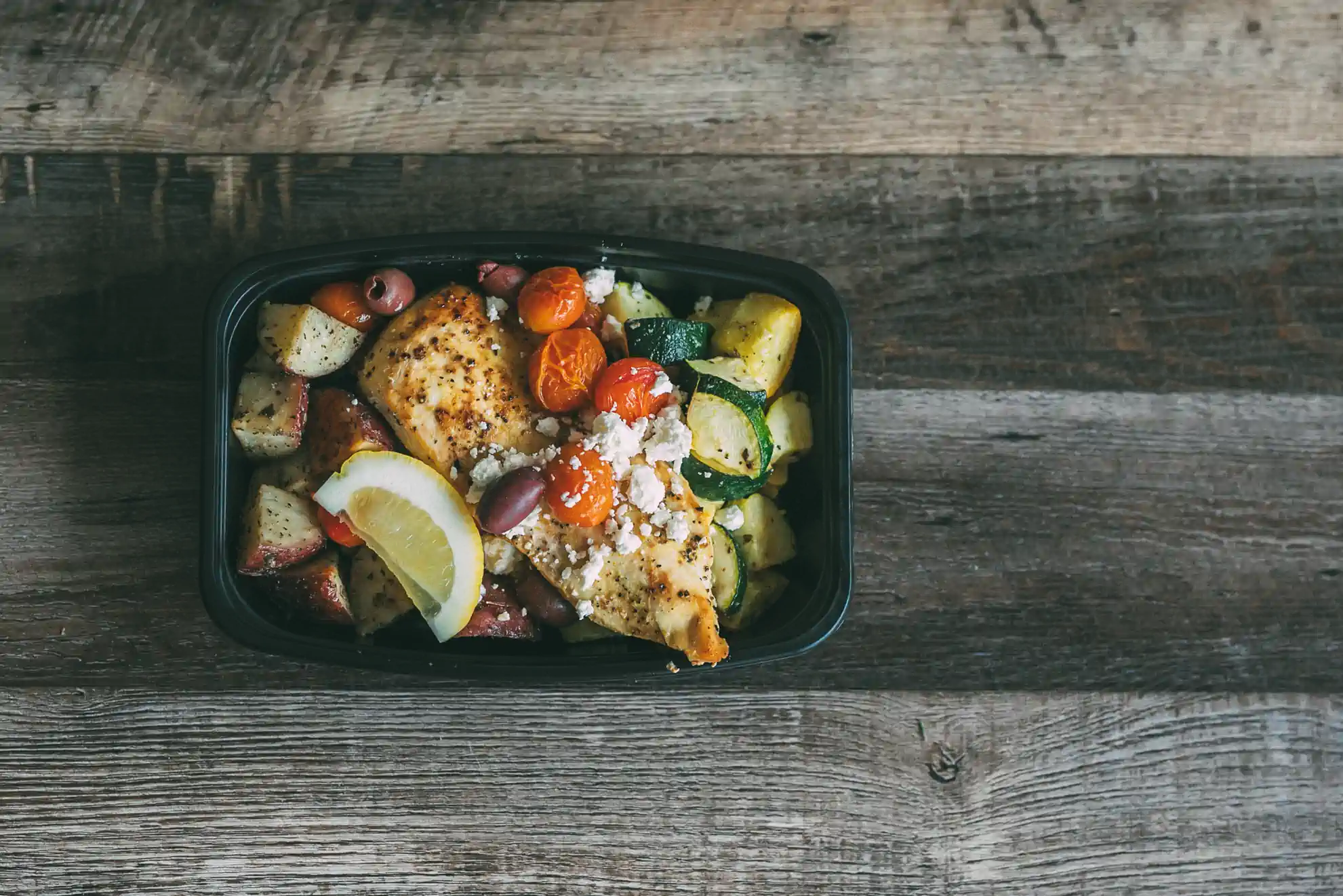 A black meal prep container from Fed by Jed contains seasoned chicken breast, roasted potatoes, zucchini, cherry tomatoes, olives, and a lemon wedge, topped with crumbled feta cheese. The container is placed on a rustic wooden surface.