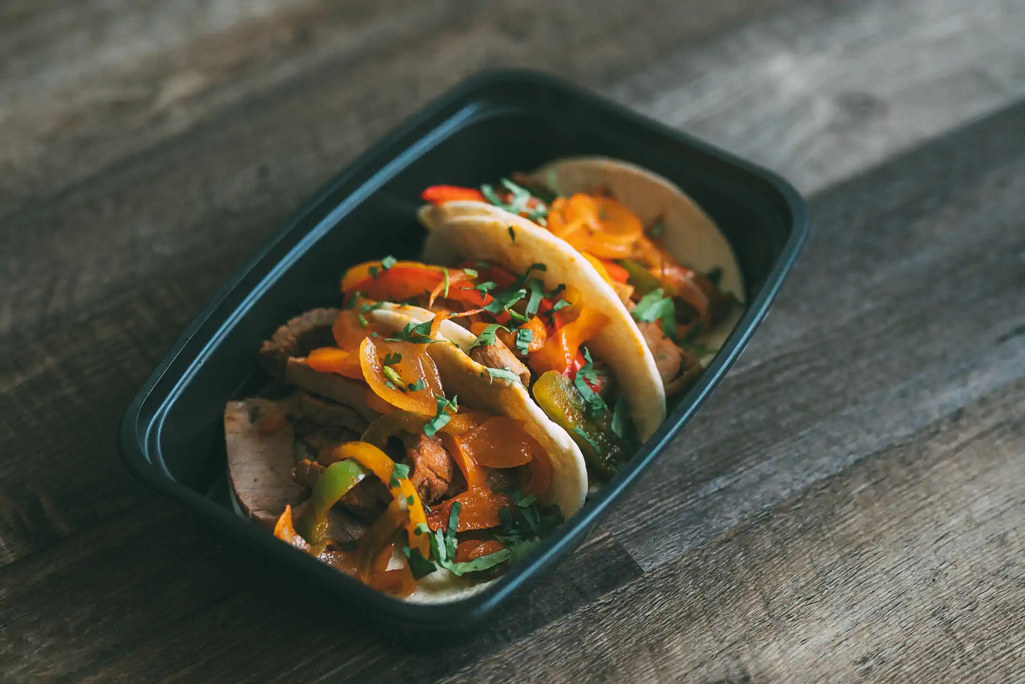 Three soft tacos filled with sautéed bell peppers, onions, and steak, garnished with fresh cilantro, served in a black meal prep container on a wooden surface.