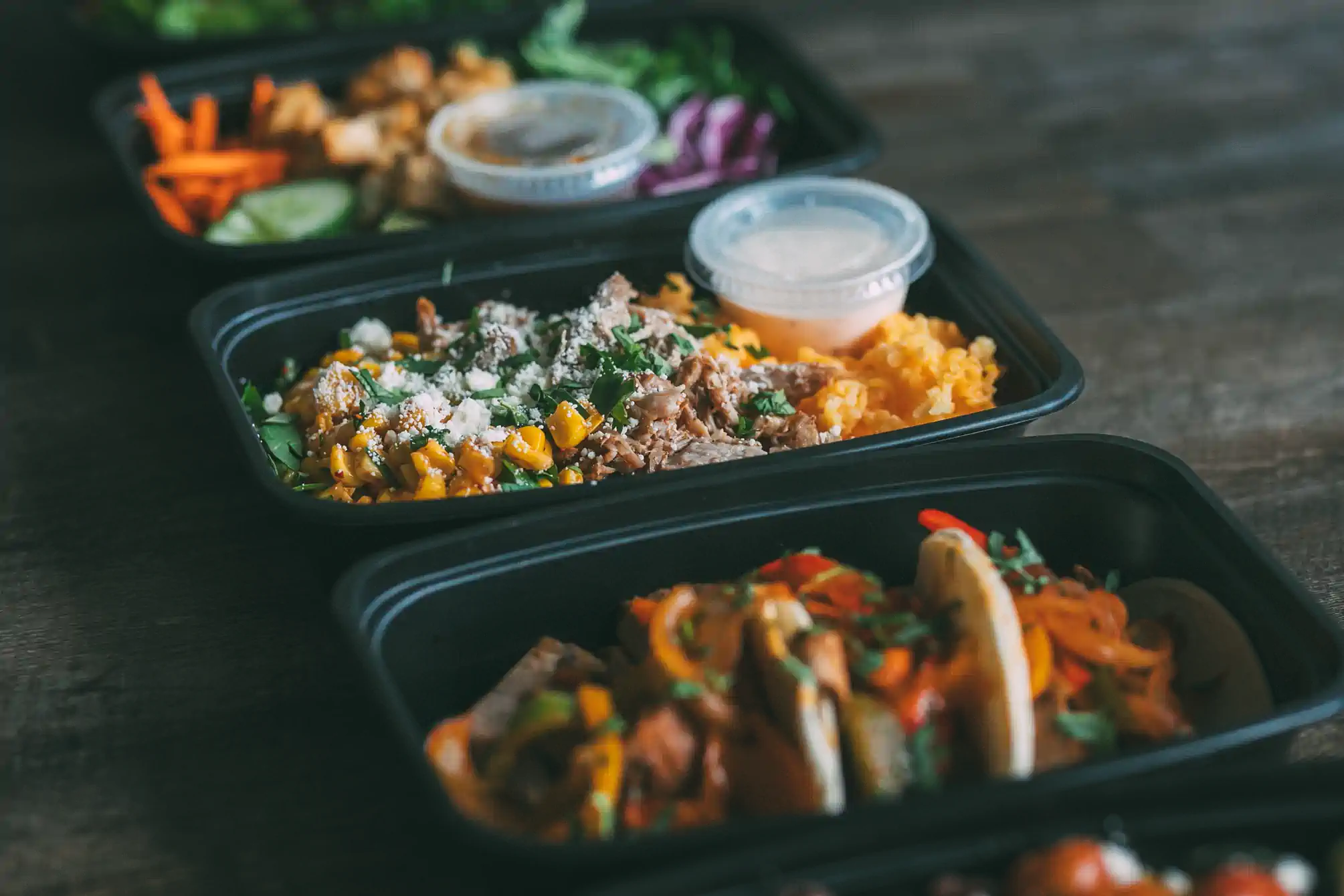 A close-up of three black meal prep containers filled with colorful, healthy food. The front container has tacos with vegetables, the middle container has a dish with corn, shredded meat, and cheese, and the back container has a variety of vegetables and a small container of sauce.