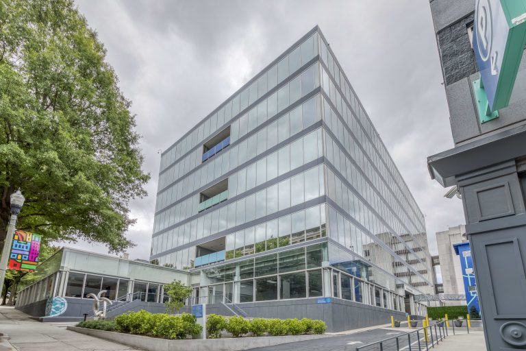 Modern glass office building with multiple floors, greenery in front, and an overcast sky.