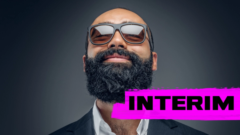 Smiling man with a full beard and sunglasses wearing a dark suit and white shirt against a gray background with the word INTERIM in bold black letters on a bright pink brushstroke.