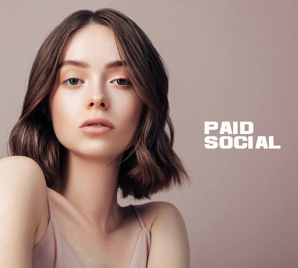 Eine Junge Frau schaut werbewirksam in die Kamera, der Hintergrund ist beige und es ist der Schriftzug “Paid Social” zu lesen, der für Werbemaßnahmen über Attitudes Eleven steht